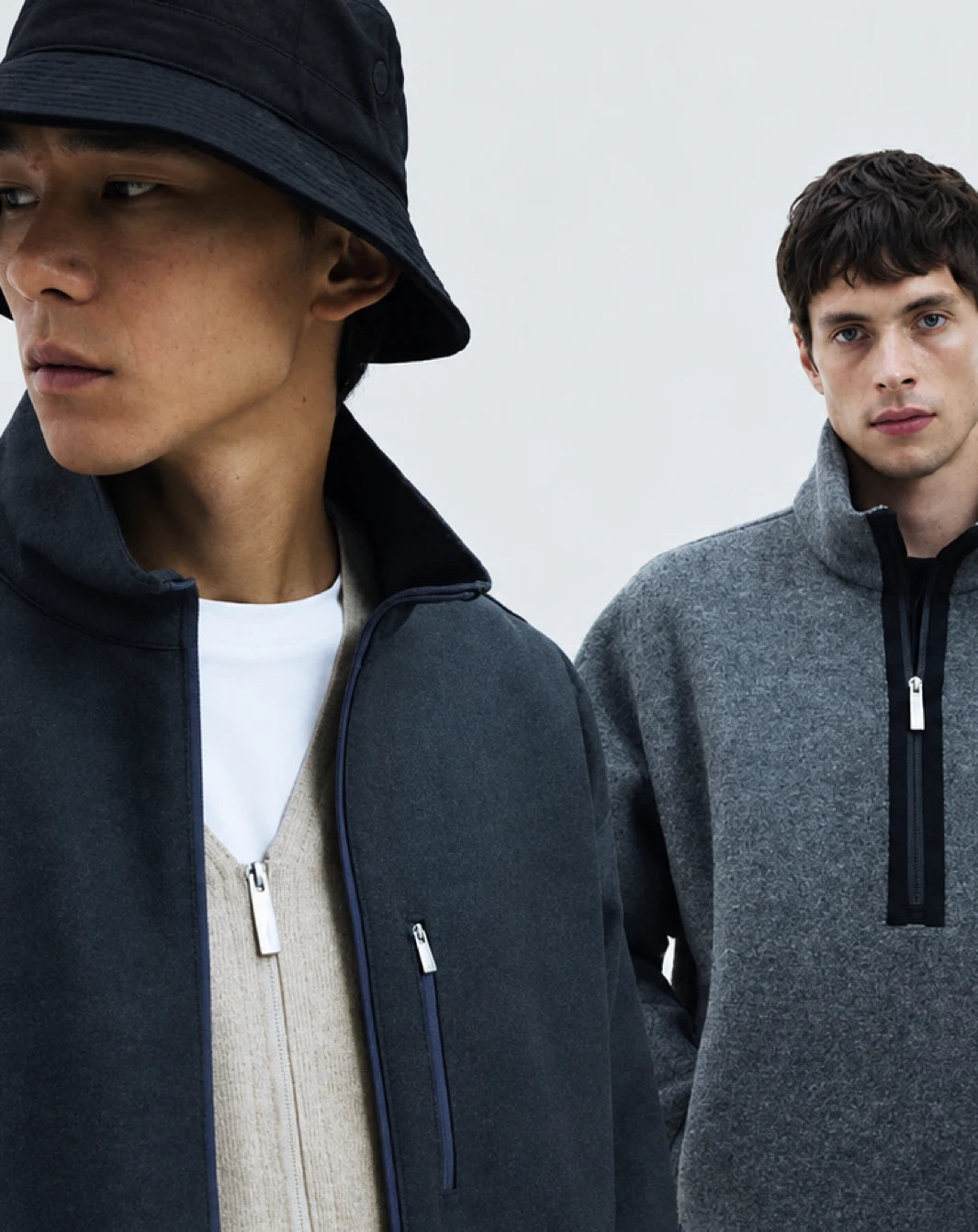 Laminar Hombre Amsterdam - Two male models wearing minimalist outerwear. The foreground model wears a black bucket hat and a dark charcoal zip-up fleece jacket over a white t-shirt and beige sweater.